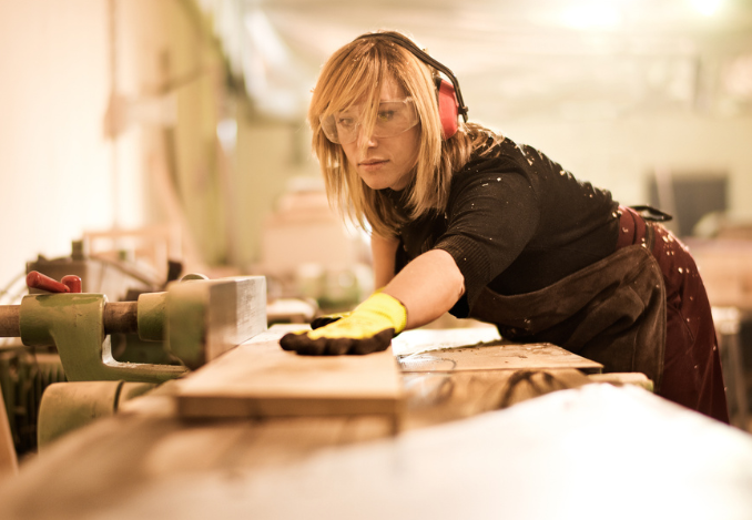 Une femme menuisier agenceur qui coupe une planche dans son atelier. Elle porte des lunettes et des gans de protection